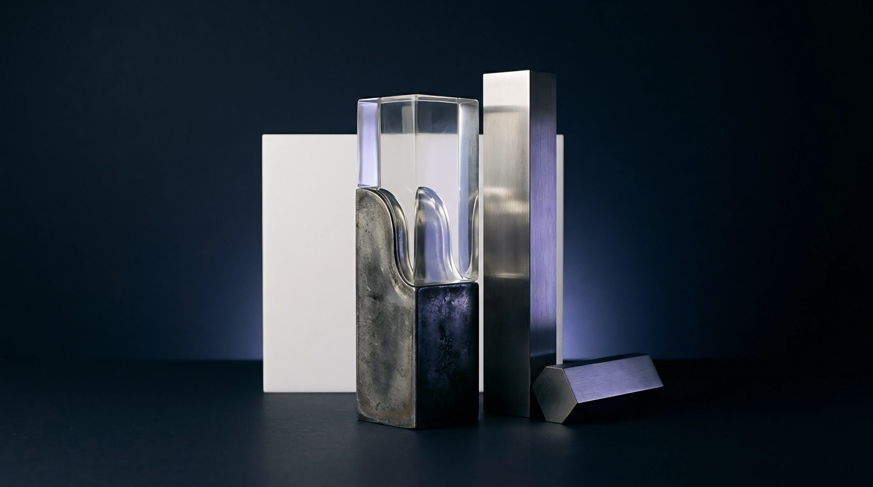 Geometric forms comprising transparent polymer with internal waves, a dark solid block, and metallic prisms stand on a dark industrial surface.