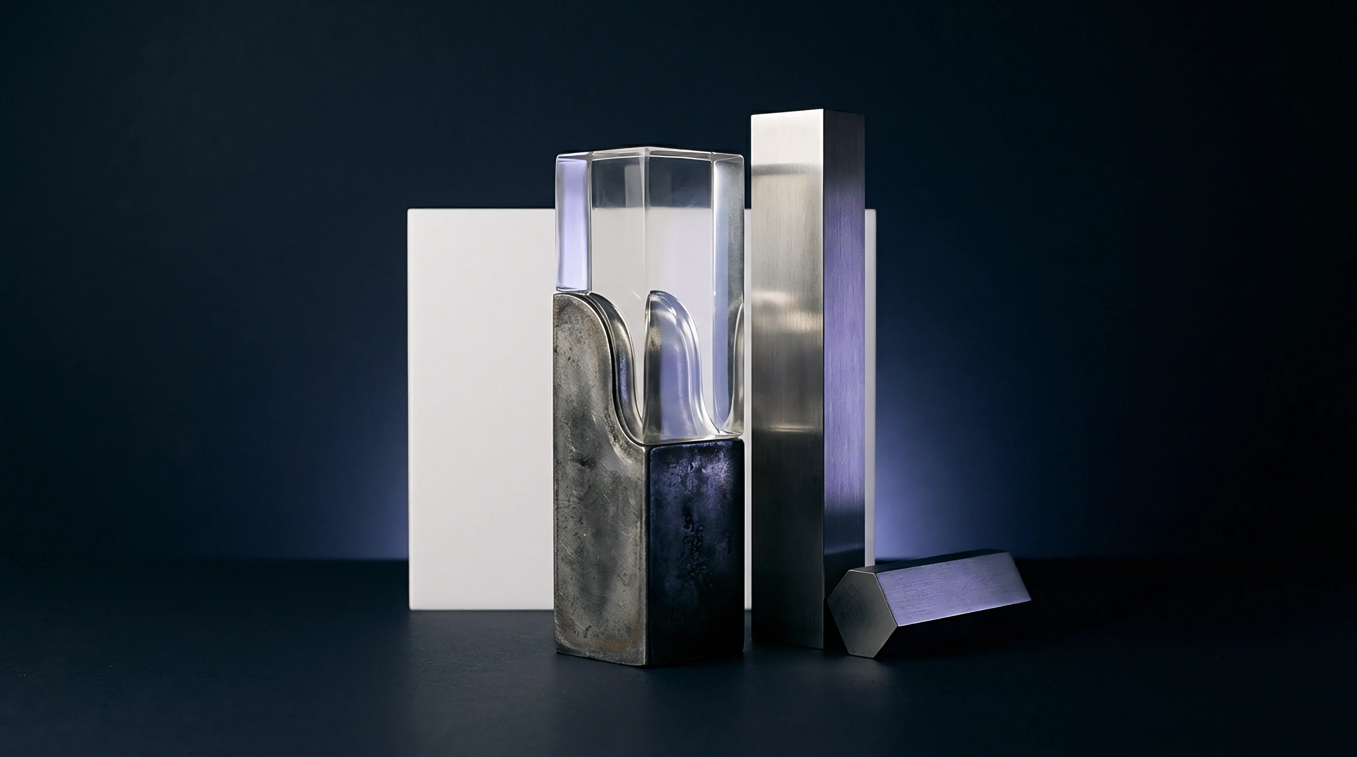 Geometric forms comprising transparent polymer with internal waves, a dark solid block, and metallic prisms stand on a dark industrial surface.