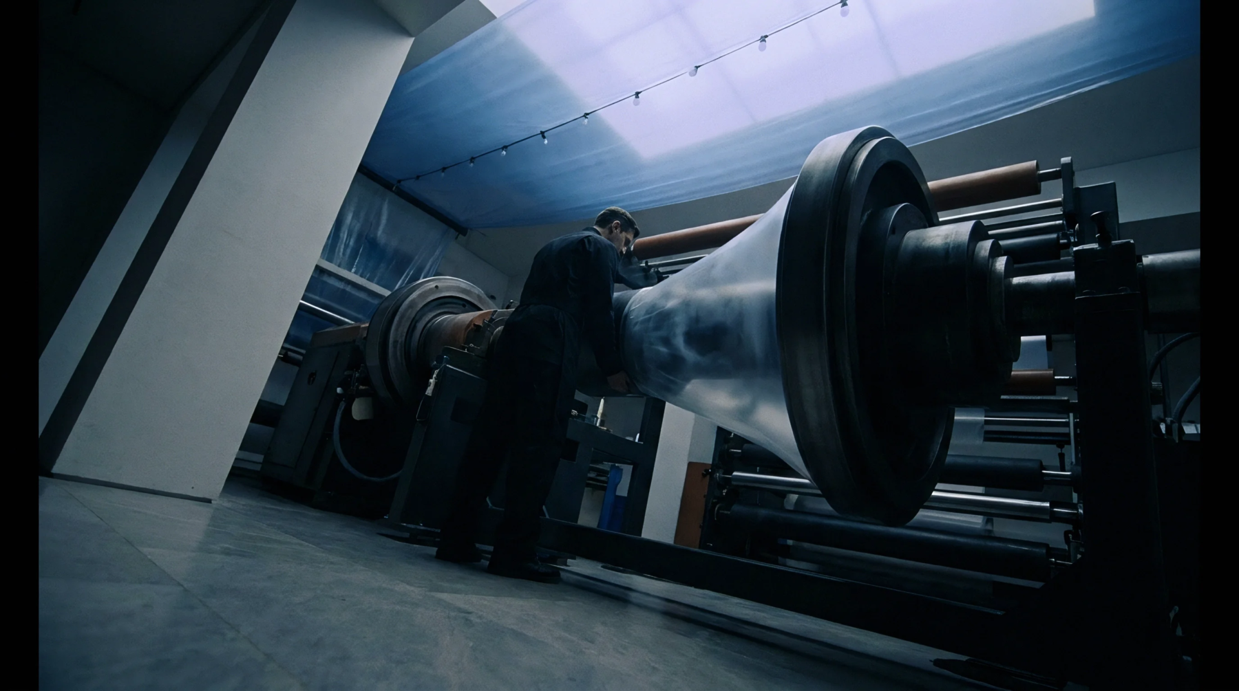 An industrial operator monitors polymer film extrusion equipment winding plastic sheeting onto a large steel roller within a production facility.