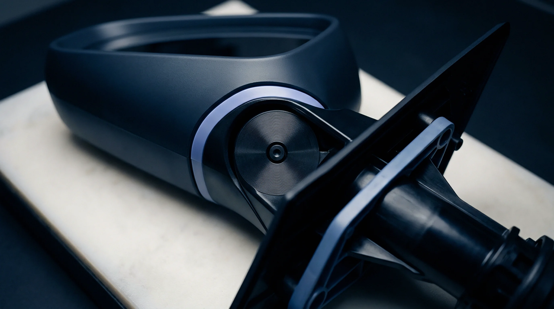 A dark engineered polymer component displays a complex molded form and an integrated pivot point, resting on a light neutral surface.